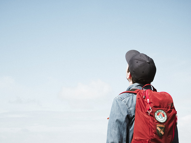 Student with red backpack and black baseball hat looking up at blue sky.