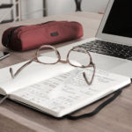 Pair of brown glasses sitting on an open notebook next to a silver laptop.