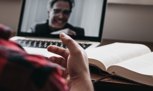 Student talking to professor through video chat on a laptop with a Bible open in front of them.