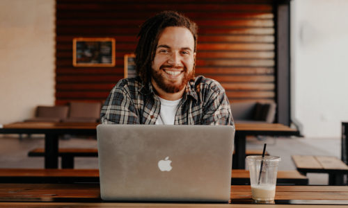 Student sitting in coffee shop using Mac laptop computer smiling at camera.