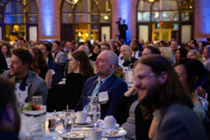 Large crowd seated at tables wearing formal clothes in an event center at night.