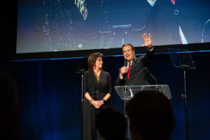 President Scott Hagan and his wife Karen speaking on stage wearing dark formal clothing in front of a projector screen.
