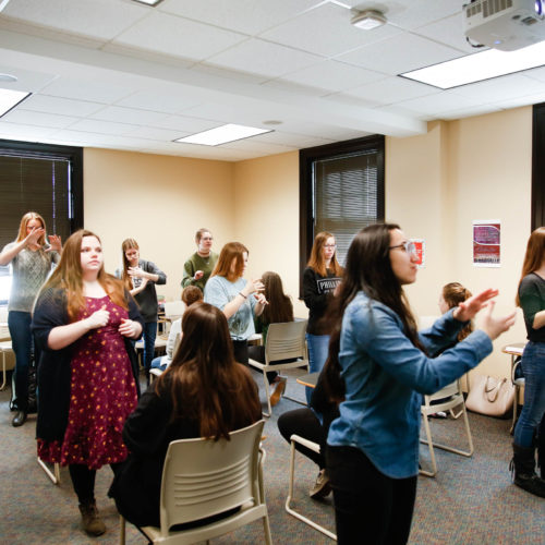 Several students stand in a classroom practicing their sign language