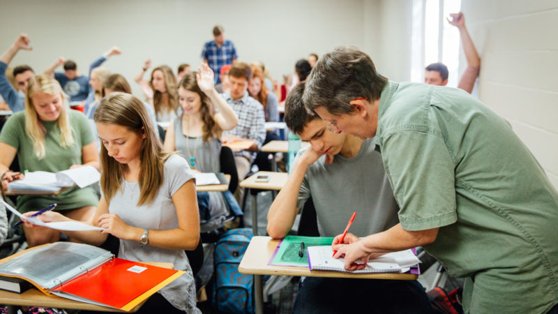 A teacher helllps a student while others work or sit with their hands raised, awaiting assistance