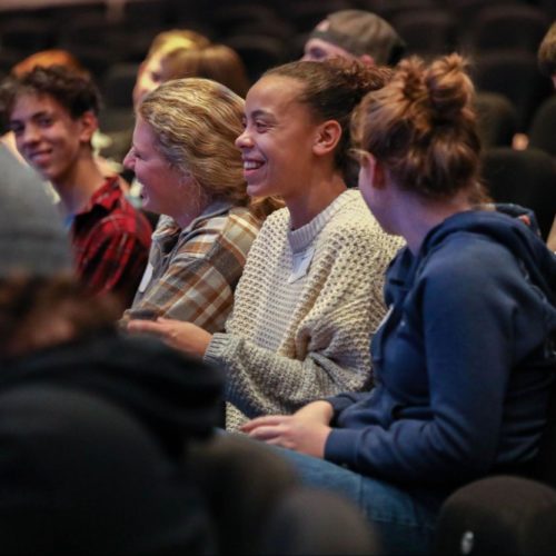 An audience smiles and watches intently in a classroom