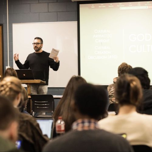 A teacher stands at the front of a classroom and speaks to his students