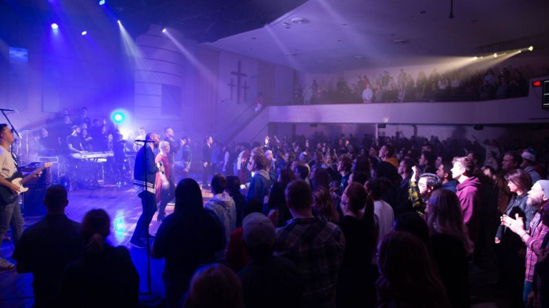 A band performs on stage for a large audience with many people on a balcony above