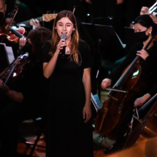 Abbie sings into a microphone with an orchestra around her