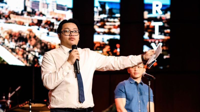 A man speaks on stage to an audience with a band behind him