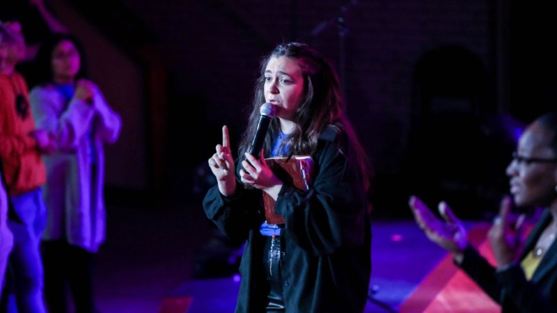 A woman stands on stage with a microphone speaking intently to an audience, a sign language interpreter can be seen at her side