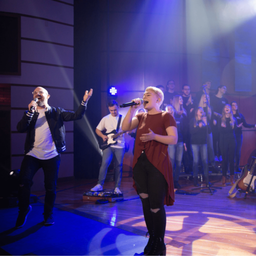 A man and woman sing together on stage with a guitarist and backing choir