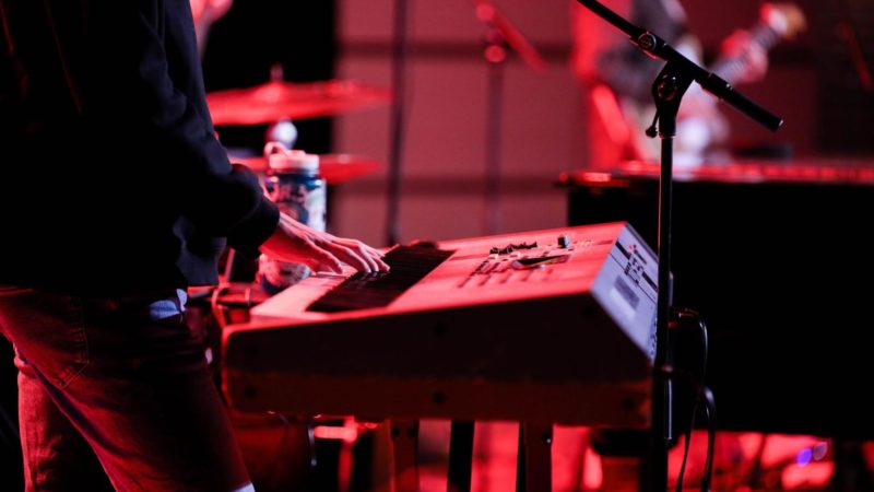 A man plays a keyboard during a live performance with his band in the background