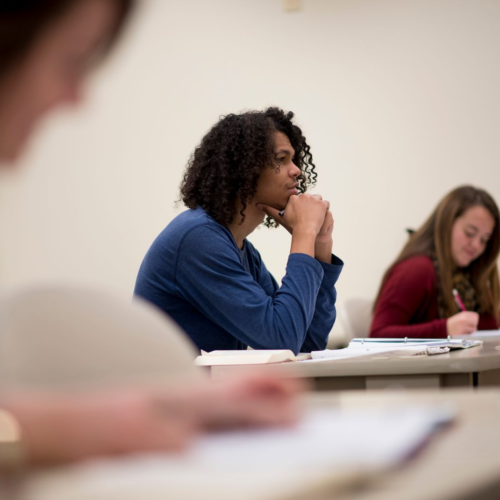 A man stares intently towards the front of a classroom while women in the foreground and background write