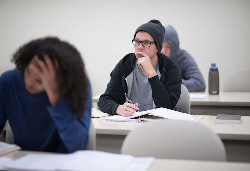 A student in a beanie looks forward with a finger on his lip, two other students working in front and behind him