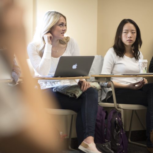 A woman sits at her desk and speaks while others around her listen