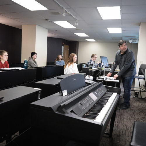In a classroom, a music teacher instructs his students on personal pianos