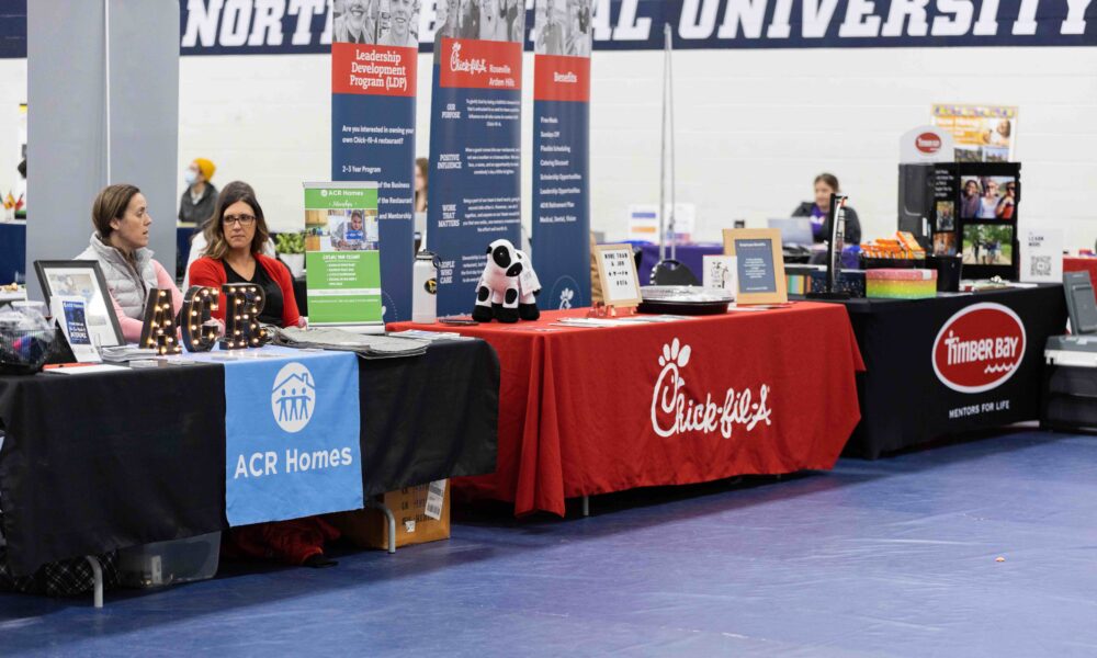 Two women sit behind an ACR Homes table, adjacent are two unattended tables for Chic-Fil-A and Timber Bay