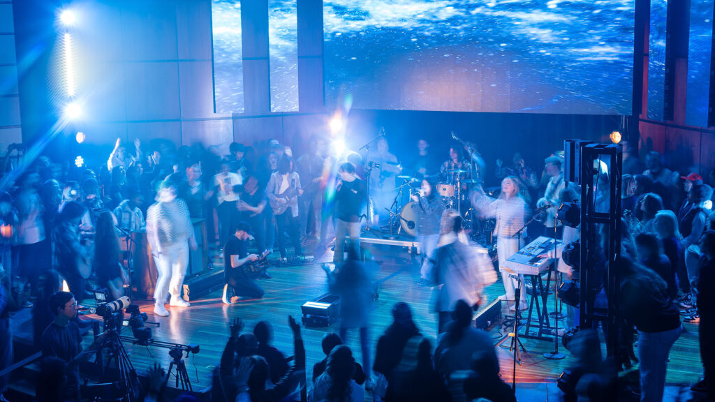 A band performs music on a stage, surrounded by an audience and various pieces of production equipment