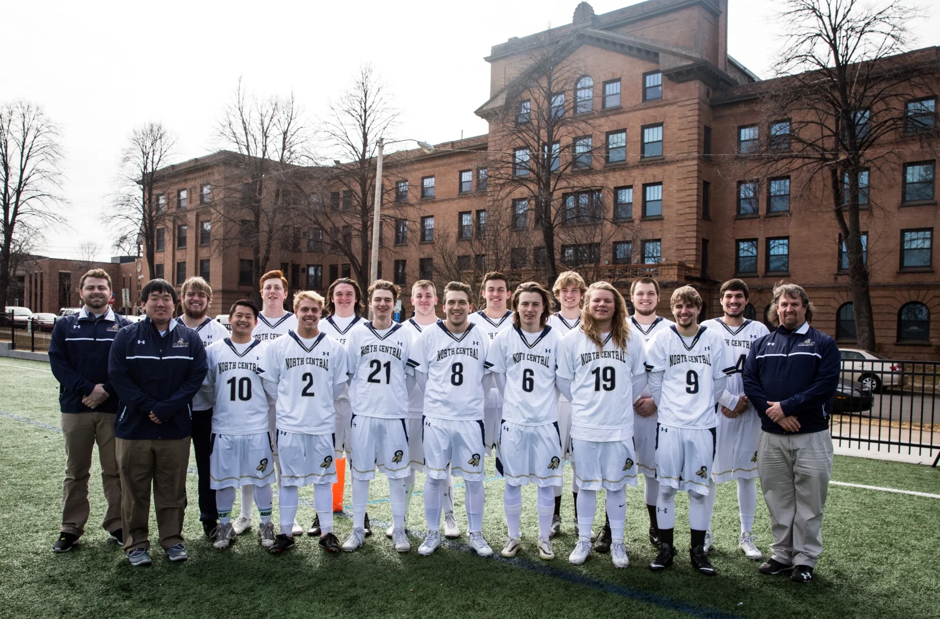 North Central University men's lacrosse team posing on the athletic field in front of a campus building