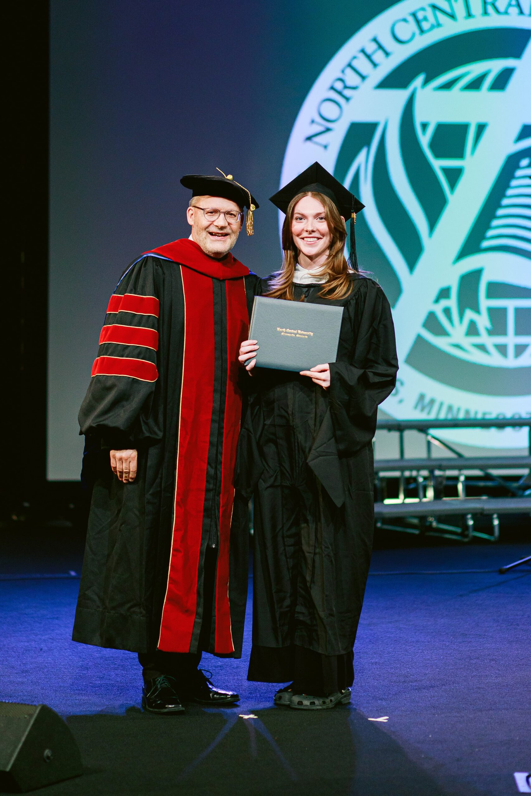 North Central University graduate receiving a diploma on stage during commencement, standing beside a faculty member in academic regalia.